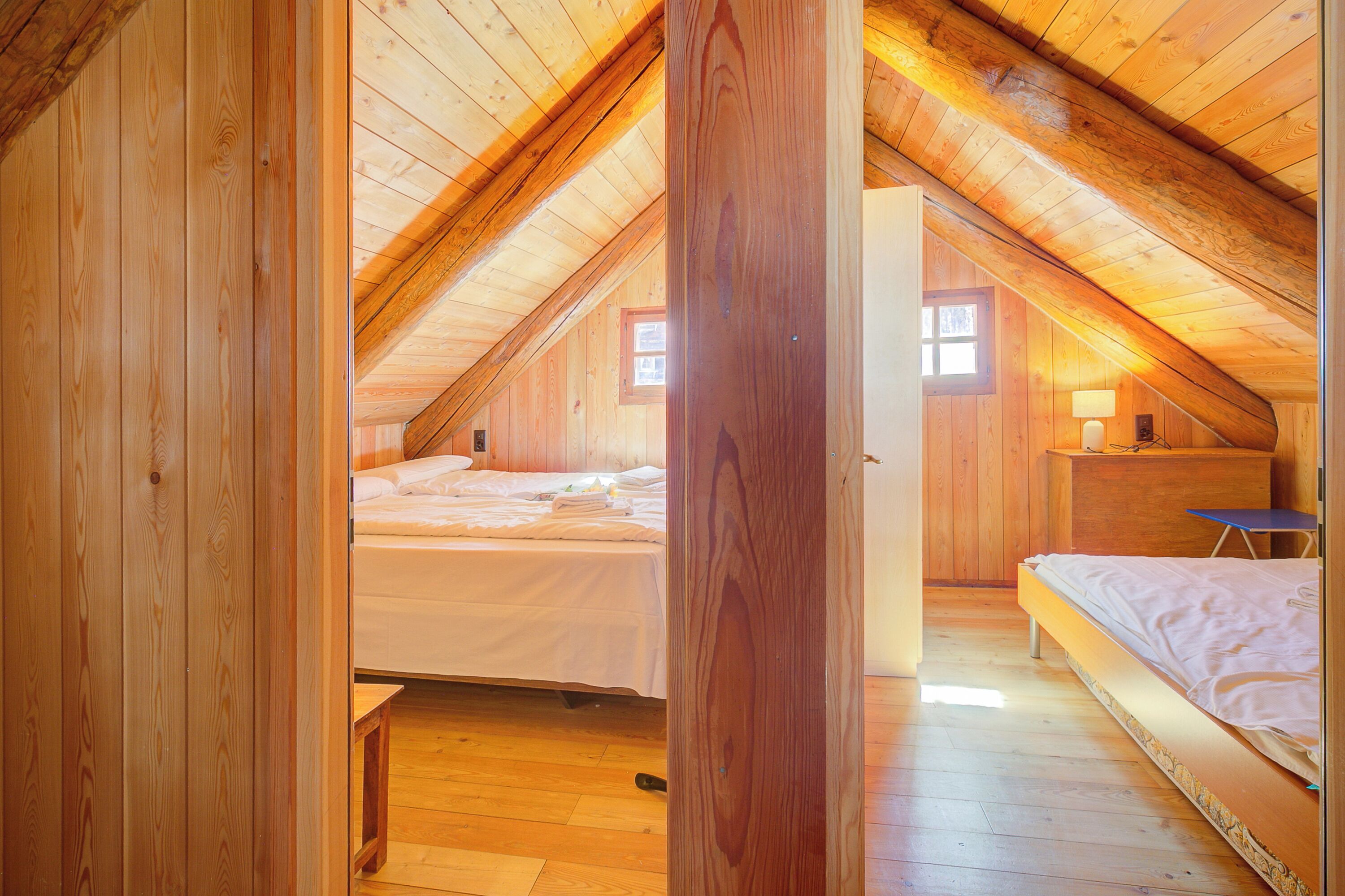 A wooden attic bedroom with two beds, a desk, and a window.