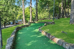 Un campo de golf con césped artificial verde, bordes de piedra y árboles al fondo.