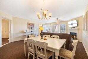 A dining area with a white table, chairs, and a chandelier. There is a fireplace with a TV mounted above it.