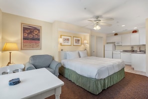 Habitación de hotel con cama, escritorio, silla, ventilador de techo, lámpara y área de cocina con microondas y refrigerador.