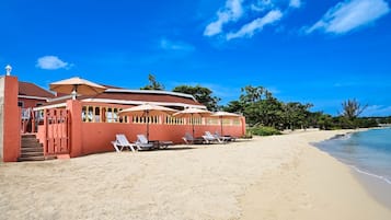 Un bâtiment situé en bord de mer, avec une façade rouge, des tables à l’extérieur protégées par des parasols et une plage de sable.