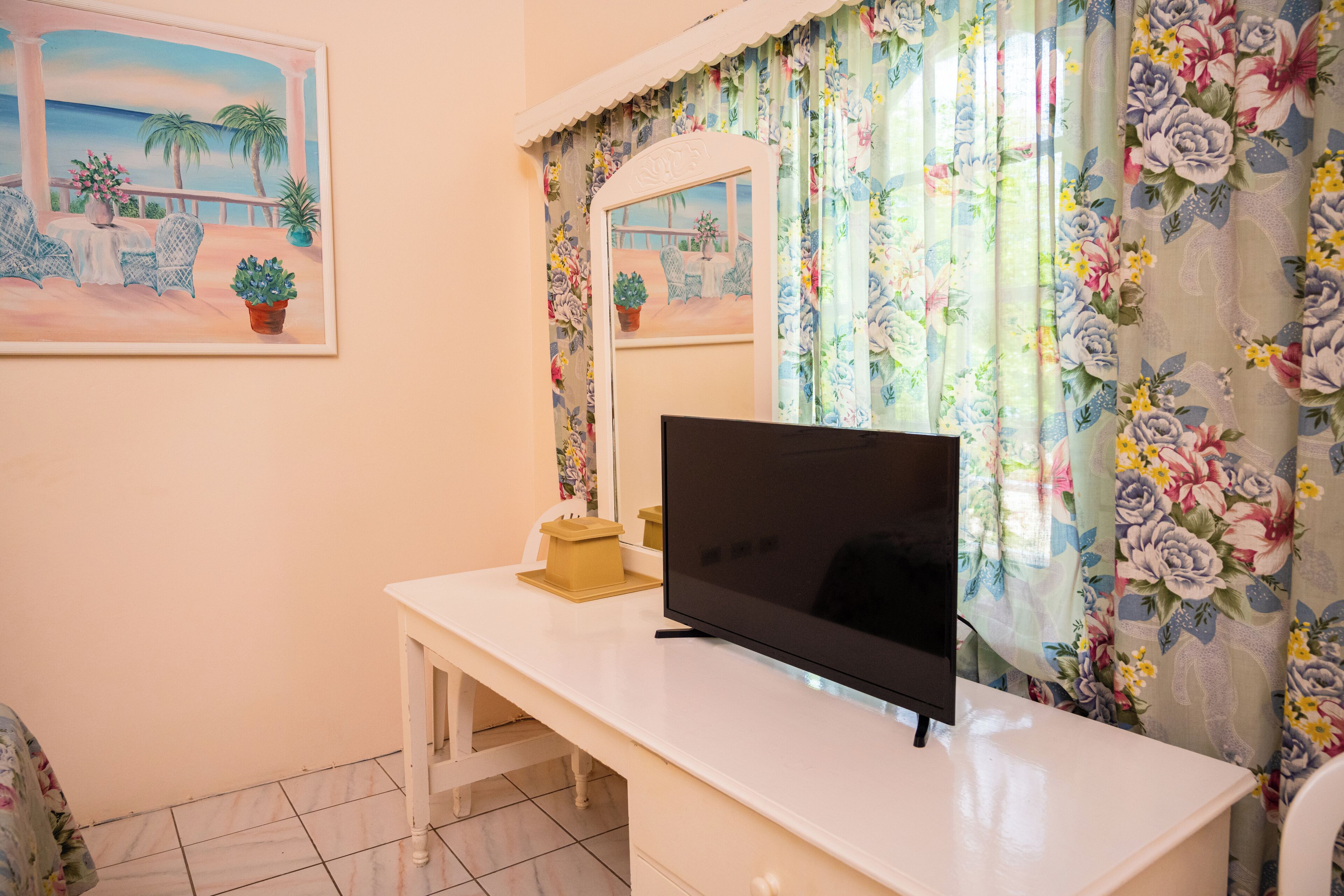 A room with a white desk, a flat-screen TV, a mirror, and floral curtains.
