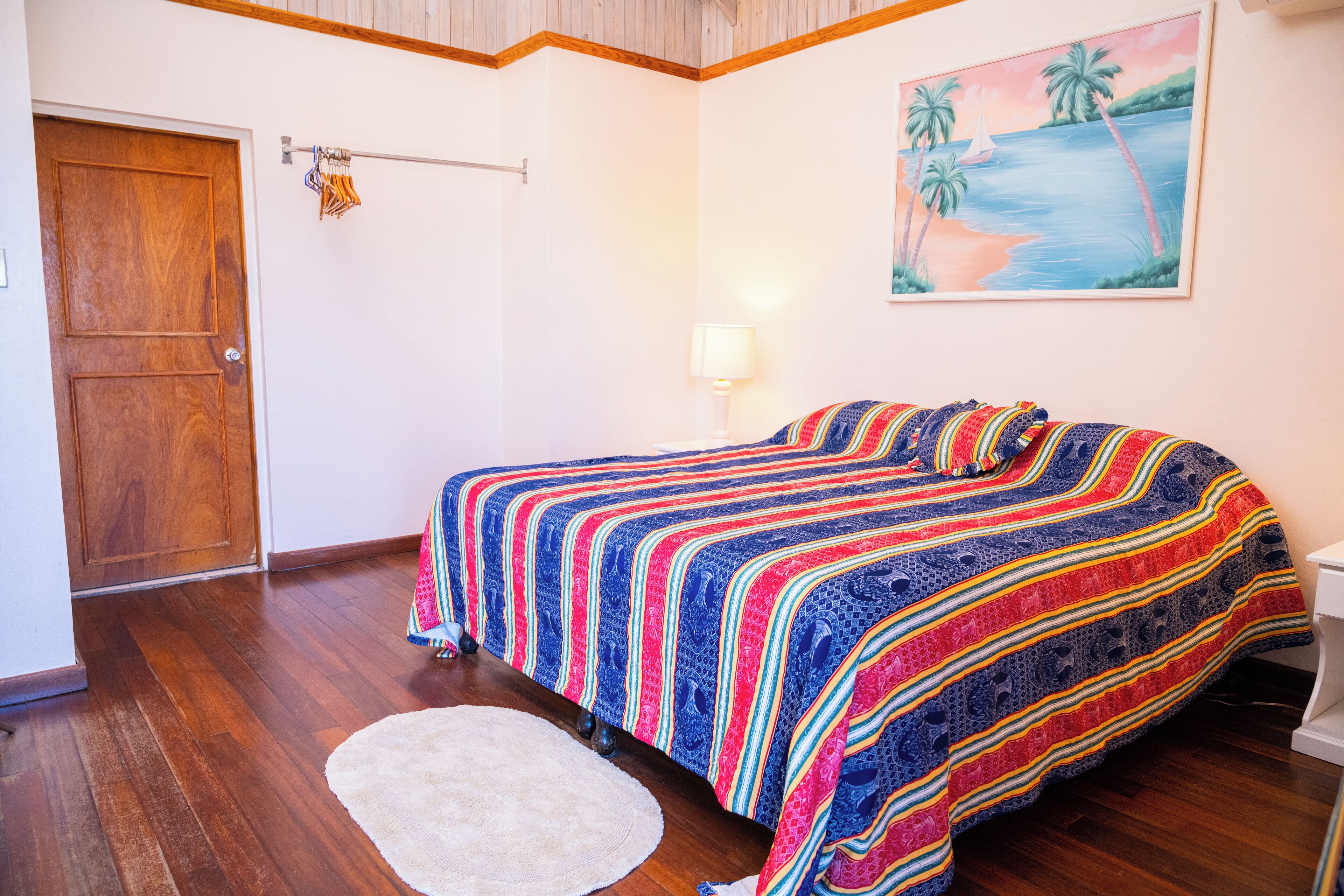 A bedroom with a wooden door, a bed with a colorful striped blanket, a white rug, and a painting of a beach scene on the wall.