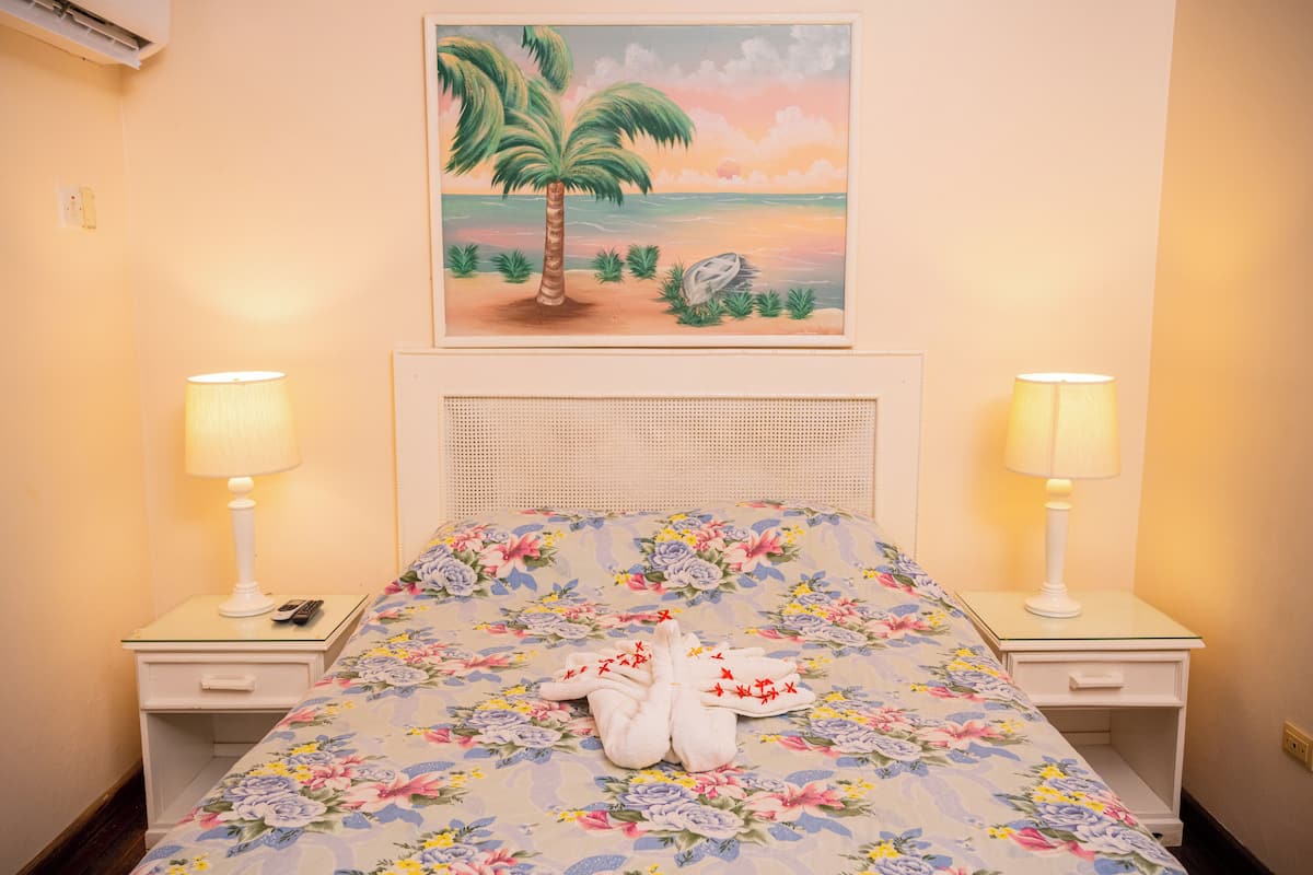 Una habitación de hotel con una cama, cubrecama floral, dos lámparas de noche, un cuadro con una playa y una mesita de noche con un control remoto.