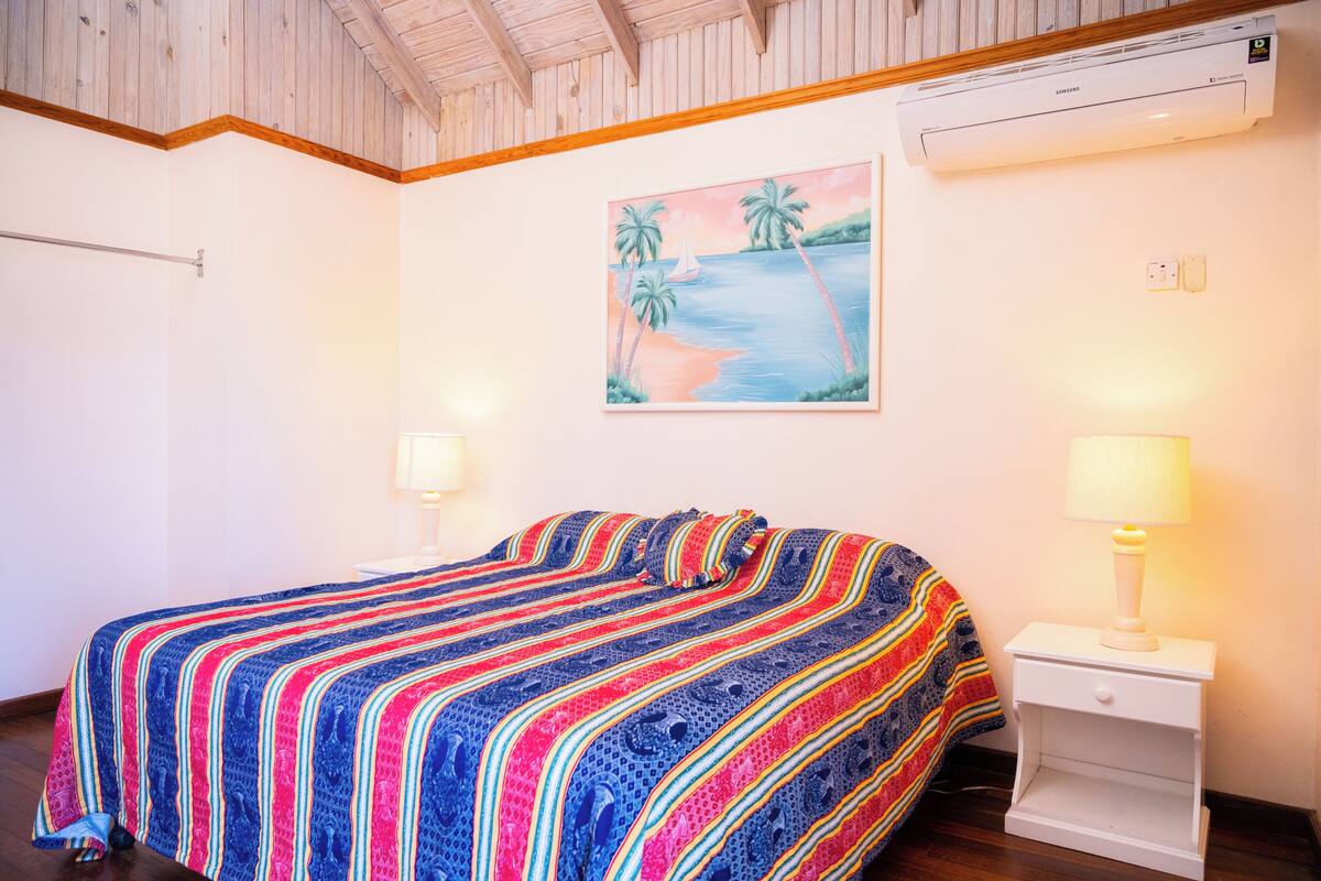 Un dormitorio con cama, una mesita de noche, aire acondicionado de pared y un cuadro con una playa.