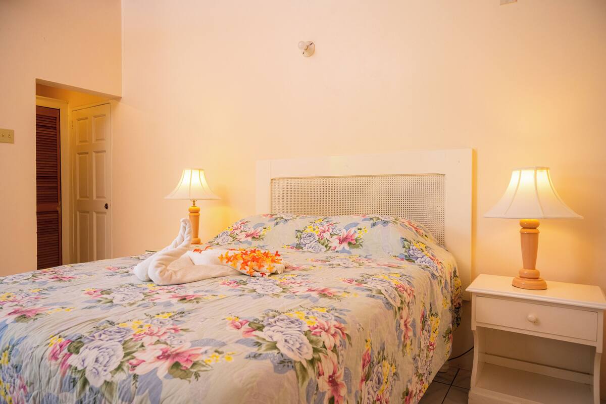 Una cama bien hecha con ropa de cama floral, dos lámparas de noche y una mesita de noche blanca.