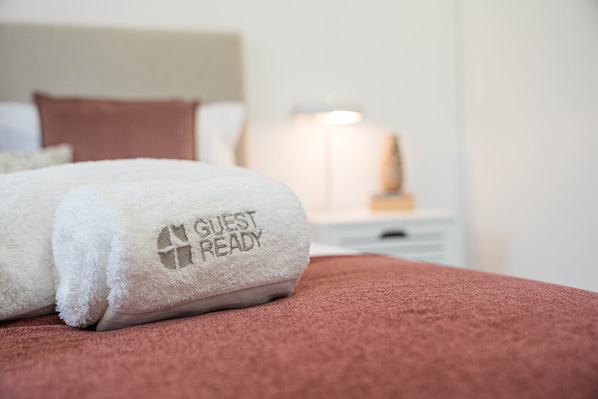 A neatly folded white towel with "GUEST READY" embroidered on it, placed on a bed with a pink blanket.