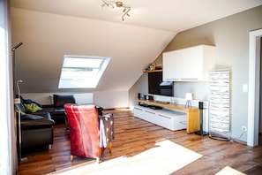 A modern loft apartment with a sloped ceiling, a large skylight, a black leather sofa, a red armchair, a wooden dining table, a built-in TV, and a white cabinet. - FeWo Siebenstern**** - 4-star vacation apartment in prime location with balcony and garage (Winterberg)