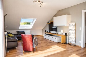 A modern loft apartment with a sloped ceiling, a dining area, a sofa, a red chair, a TV, and a kitchen area. - Siebenstern**** Vacation Rental - 4-star vacation rental with high-speed Wi-Fi, balcony & (Winterberg)