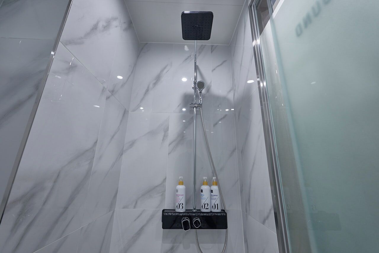 A modern bathroom with a glass shower enclosure, marble walls, and a black shelf holding three bottles.