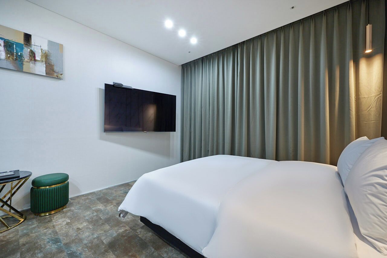 A modern hotel room with a large bed, a flat-screen TV mounted on the wall, a small green ottoman, and a painting on the wall.