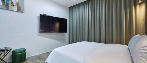 A modern hotel room with a large bed, a flat-screen TV mounted on the wall, a small green ottoman, and a painting on the wall.
