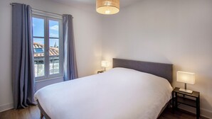 Ein Schlafzimmer mit einem großen Bett, zwei Nachttischen mit Lampen, einem Fenster mit Vorhängen und Blick auf Gebäude.