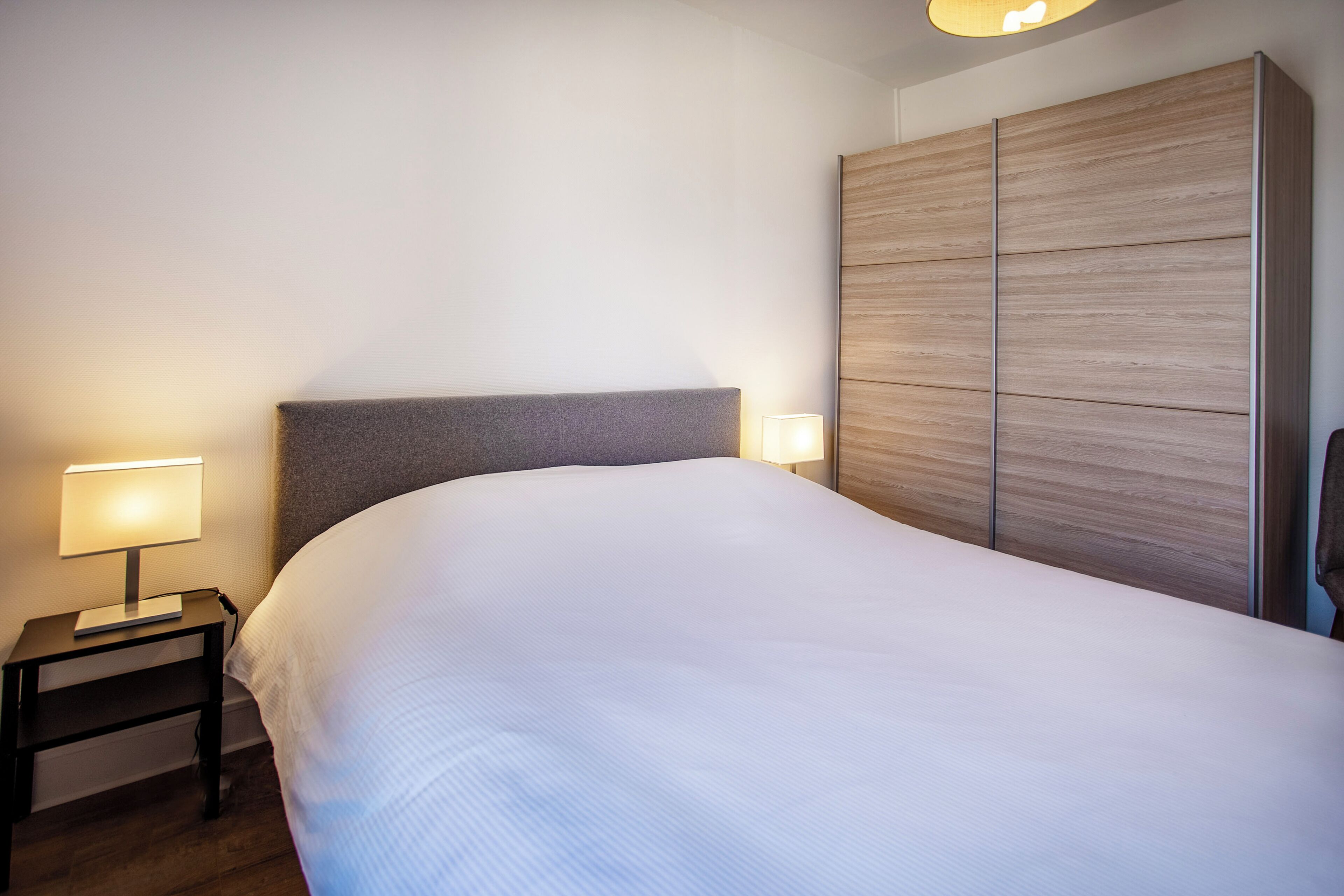 Una cama bien hecha con cabecera gris, una lámpara de noche y un armario de madera.