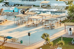 An outdoor pool area with multiple seating arrangements, including sun loungers and tables, surrounded by umbrellas and shaded by pergolas. - Boomerang Village (Manduria)