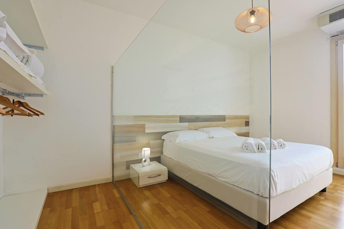 Une chambre moderne avec un plancher en bois, un lit recouvert de draps blancs, une table de chevet avec une lampe et un luminaire suspendu.