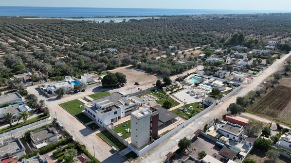 An aerial view of a residential area with modern houses, a large open space with a pool, and a view of the sea. - Boomerang Village (Manduria)