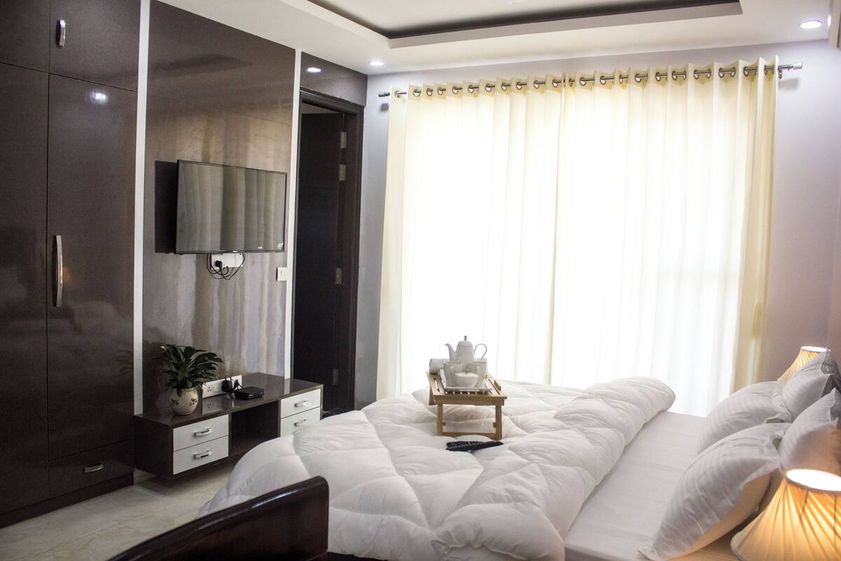 A bedroom with a bed, a TV mounted on the wall, a bedside table with a lamp, and a window with curtains.