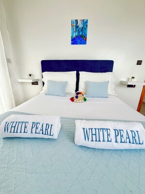 A neatly made bed with two pillows labeled 'WHITE PEARL,' a blue headboard, a small painting on the wall, and a bedside table with a plant.