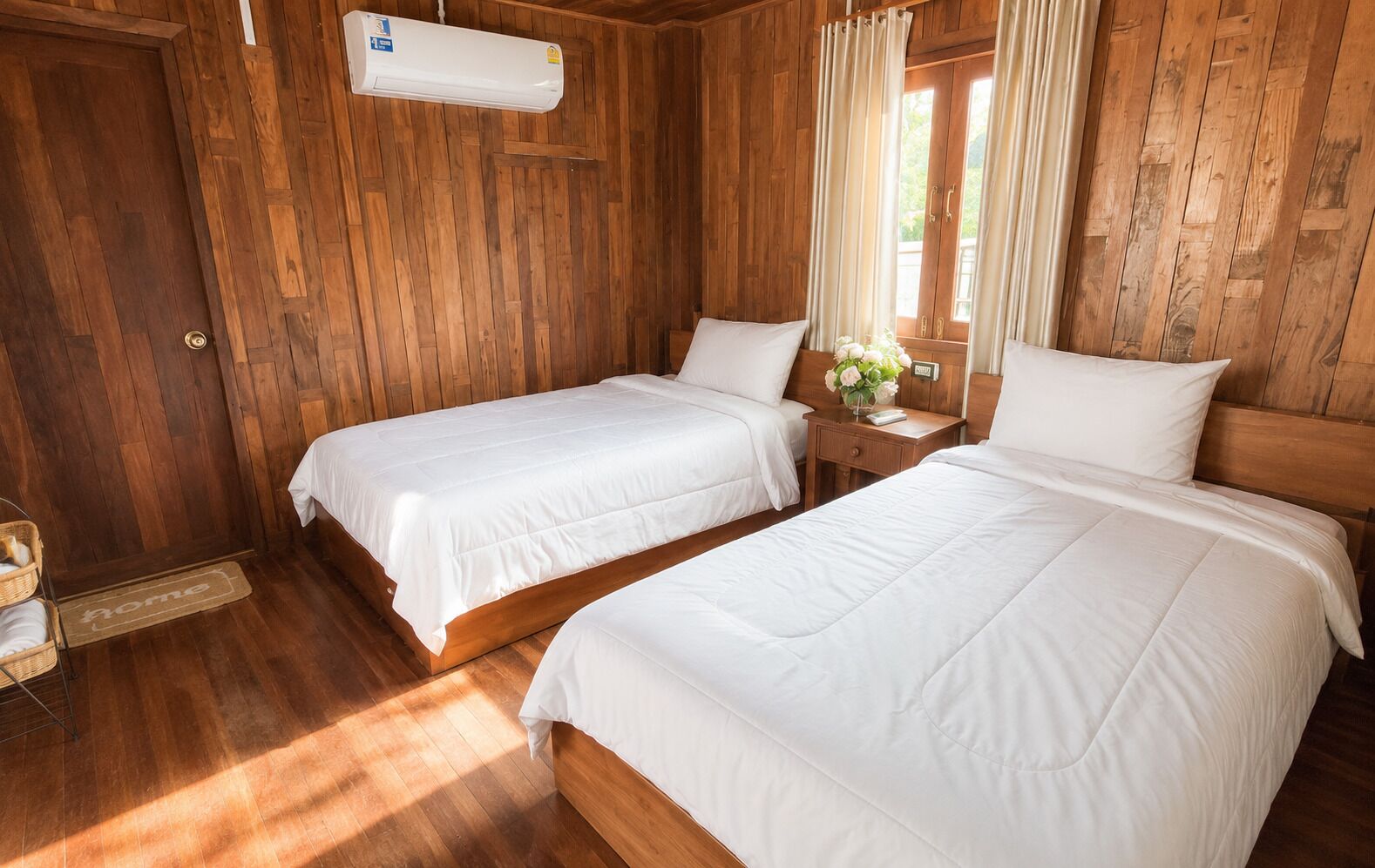 Een kamer met twee bedden, houten wanden, een raam met gordijnen, een nachtkastje met een vaas en een deur met deurmat.