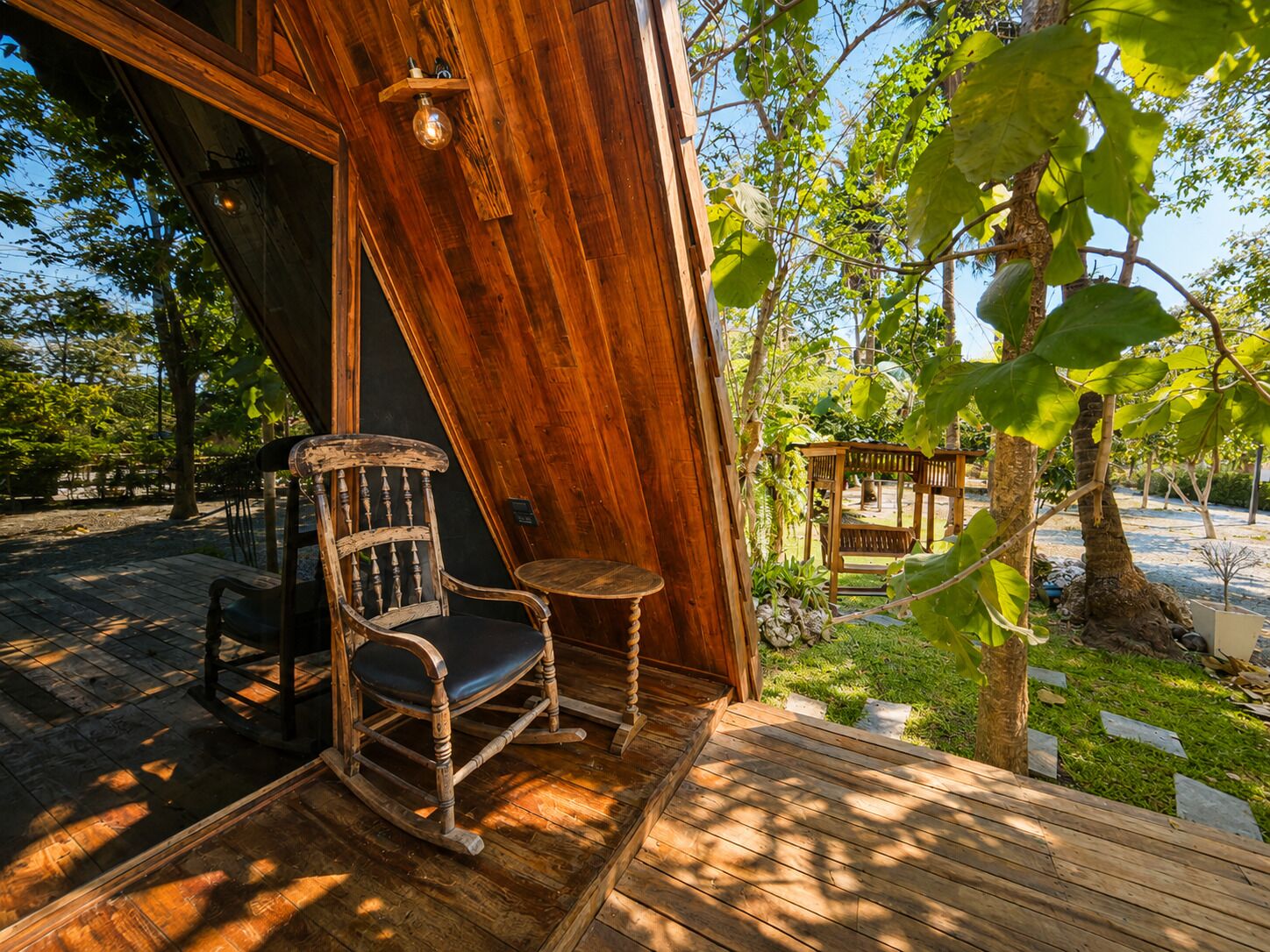 A wooden deck with a rocking chair and a small table, surrounded by greenery and trees.