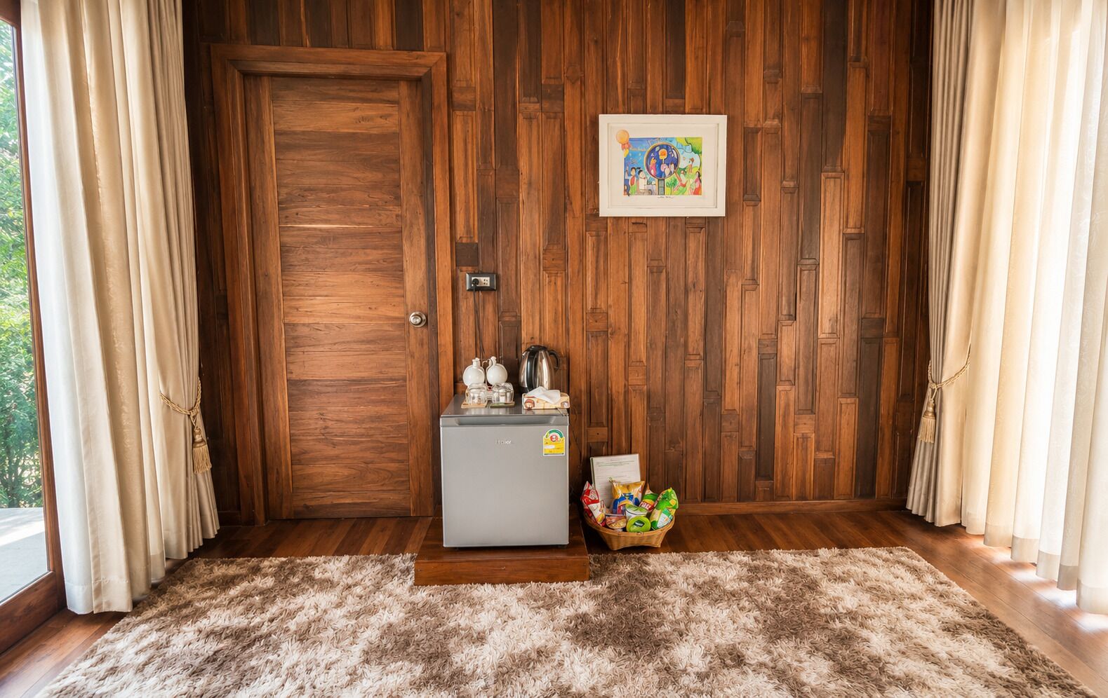 A room with a wooden door, a small refrigerator, a basket of snacks, and a framed picture on the wall.