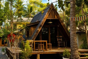 A wooden cabin with a sloped roof, a deck with seating, and a view of surrounding greenery. - Punnattha House by Peema Villa (U Thong)