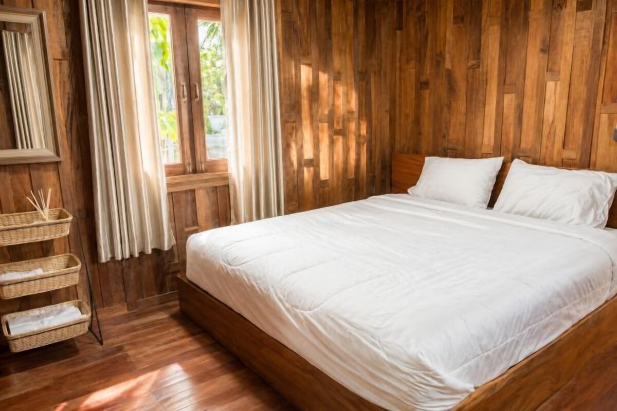A wooden-paneled room with a large bed, a window with curtains, and a bedside table with a basket.