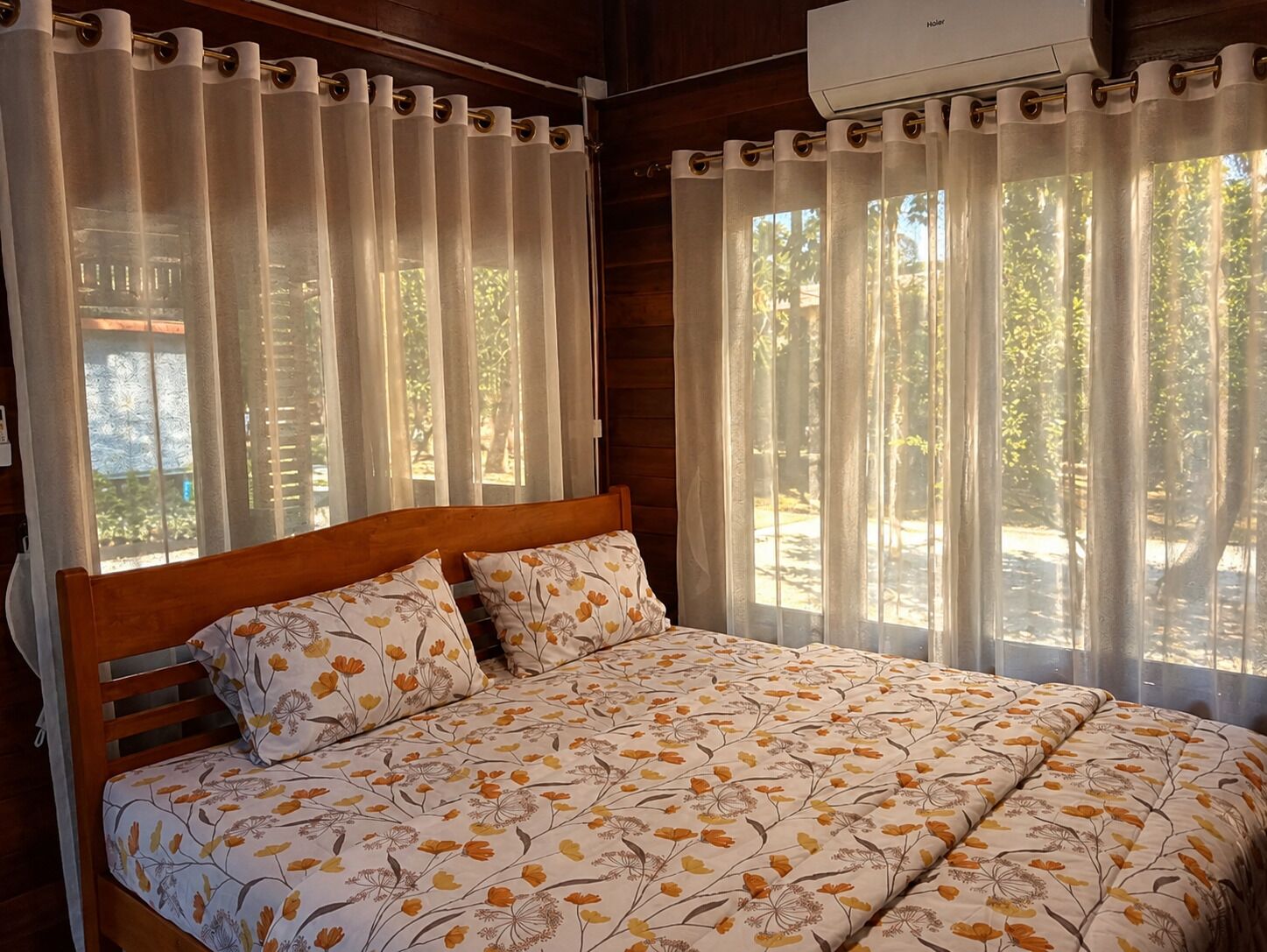 Ein Schlafzimmer mit einem hölzernen Bett, floralem Bettbezug und Blick auf Bäume durch durchsichtige Vorhänge.