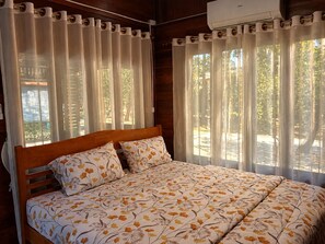 Un dormitorio con una cama de madera, ropa de cama floral y vistas a árboles a través de cortinas translúcidas.