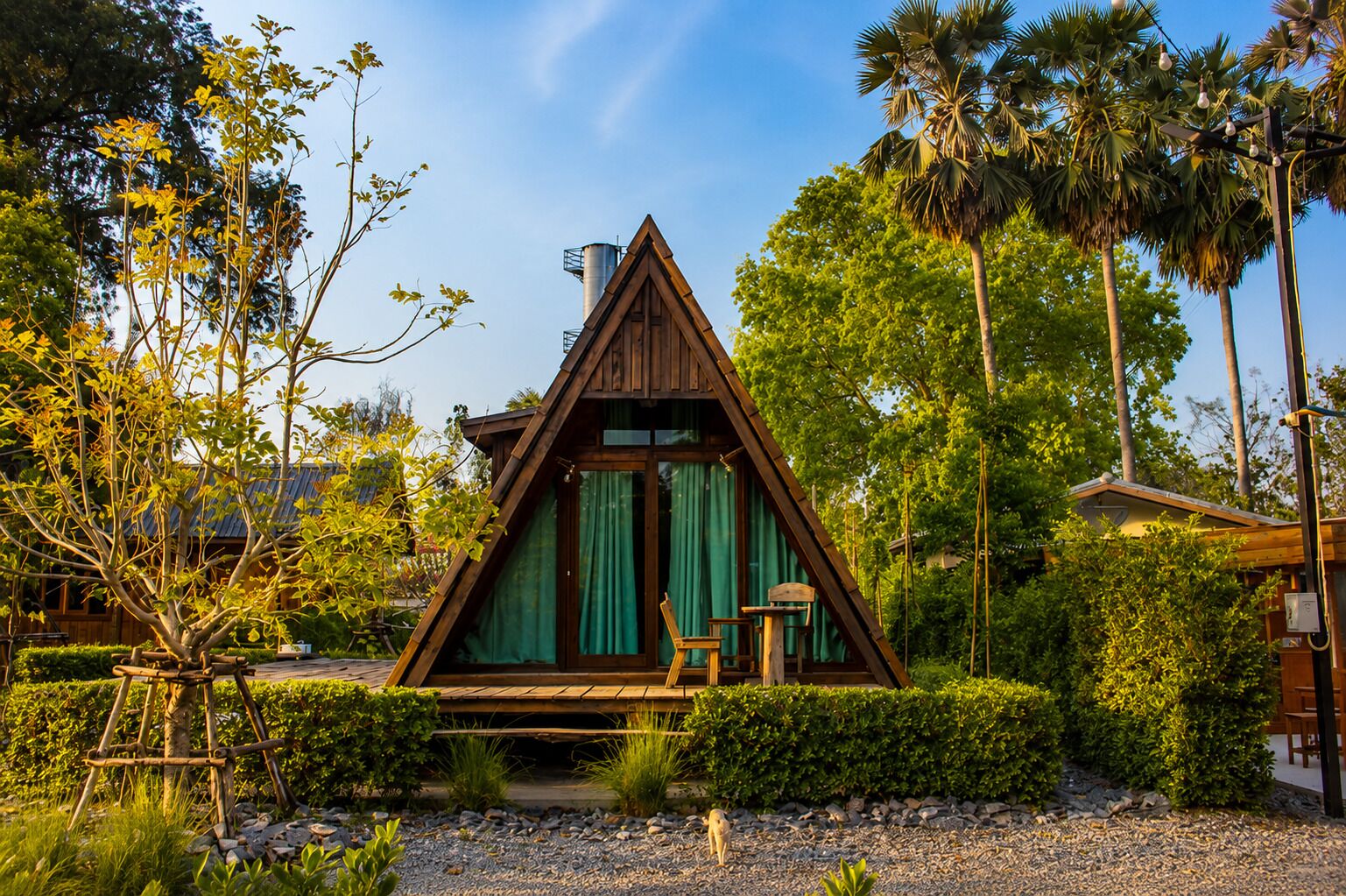 Een houten A-frame vakantiehuisje met een overdekte veranda, omringd door weelderige begroeiing en palmbomen.