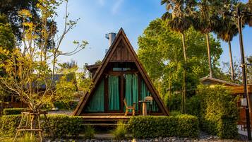 Een houten A-frame vakantiehuisje met een overdekte veranda, omringd door weelderige begroeiing en palmbomen.