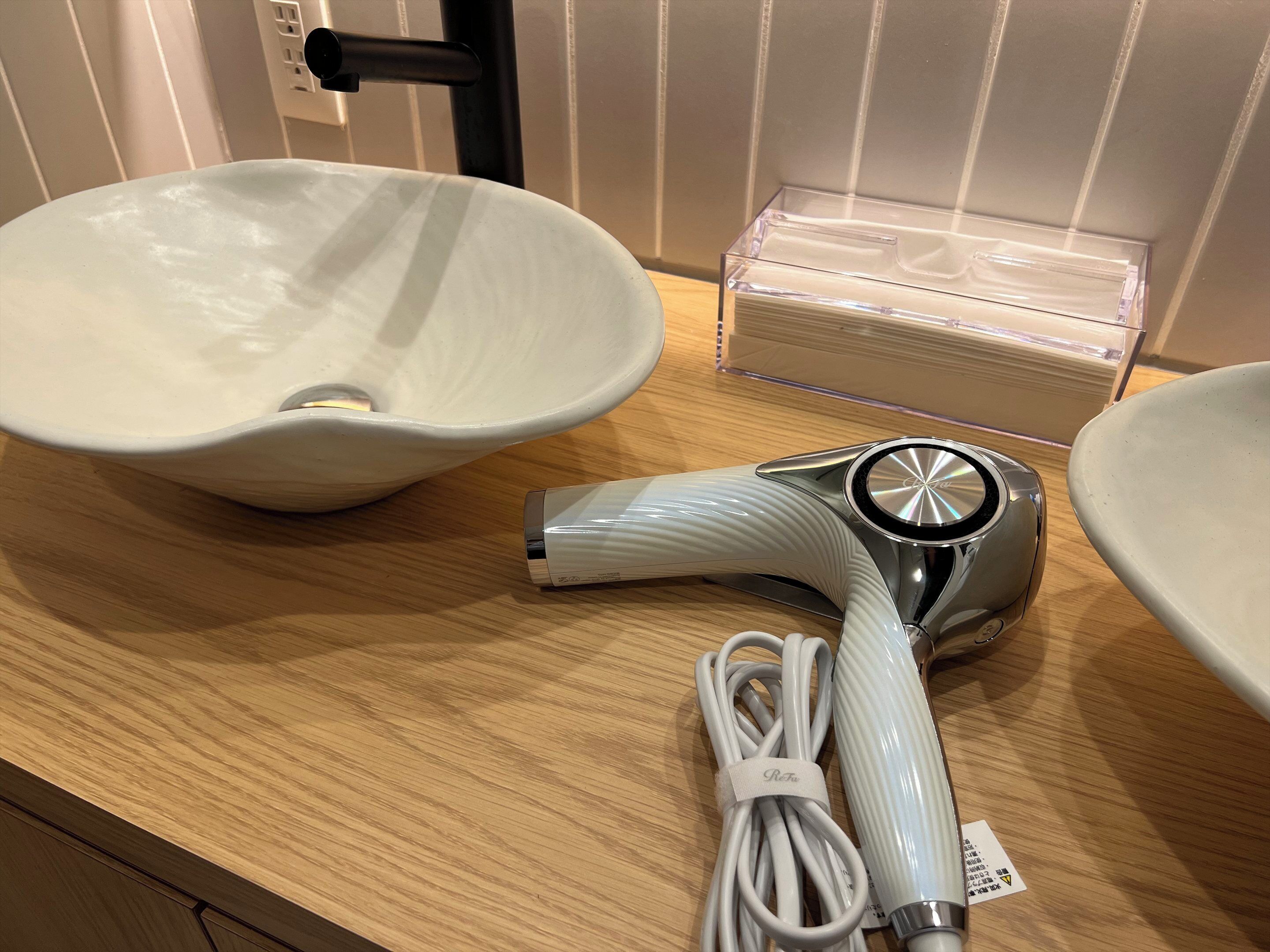 A bathroom counter with a white ceramic sink, a hairdryer, and a clear plastic storage box.