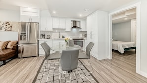 A modern kitchen with white cabinets, a stainless steel refrigerator, and a dining area with a glass table and grey chairs. - 2 bedrooms one bath walk out basement (Calgary)