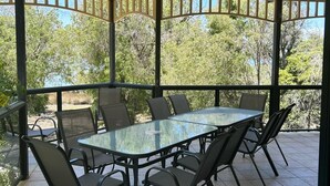 A covered patio area with a glass-top table and chairs, surrounded by a railing with a view of trees and a clear sky. - Mini Retreat Estuary 
For families (Bouvard)
