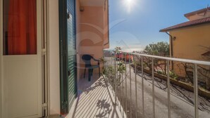Un balcón con una silla, baranda metálica y vista a una calle con coches aparcados y edificios.