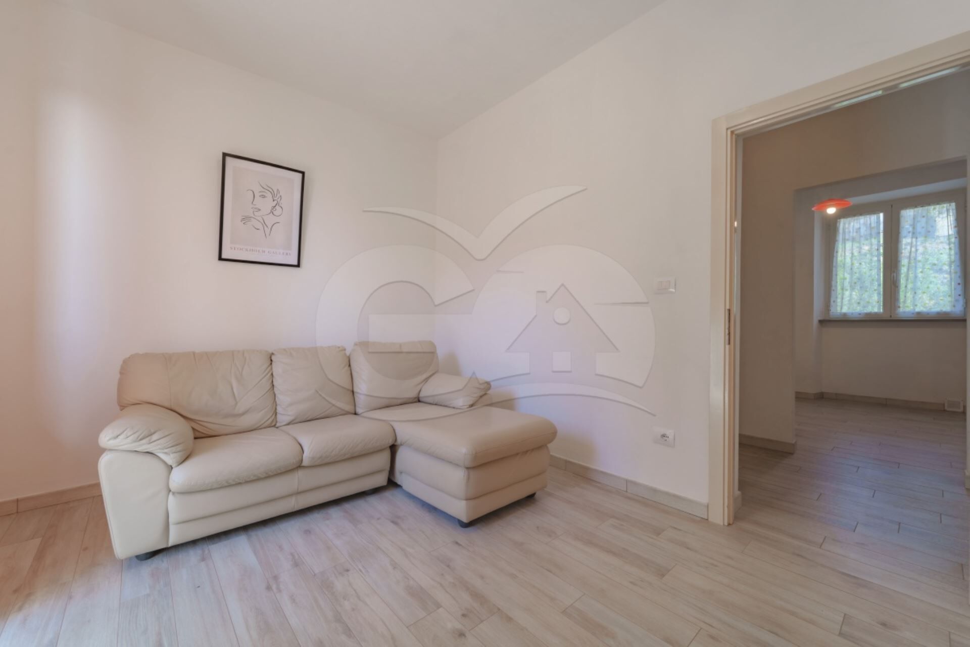 A minimalist living room with a beige sectional sofa, a framed picture on the wall, and a large window providing natural light.