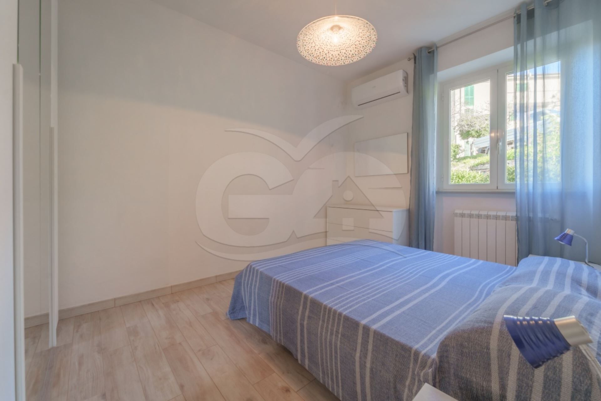 A bedroom with a bed, a window with curtains, a radiator, and a ceiling light fixture.