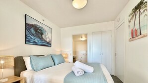 Ein ordentlich eingerichtetes Schlafzimmer mit Bett, Nachttisch und einem Gemälde einer Welle.