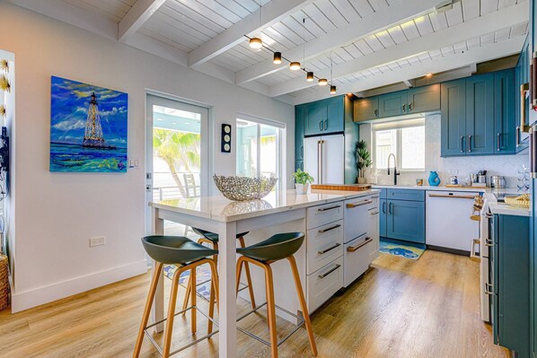 A modern kitchen with blue cabinets, a central island, bar stools, and a painting of a sailboat. - Sunset Serenity – Open Water Paradise | Dockage, Sunset Views, Dog Friendly & Minutes to Key West (Summerland Key)