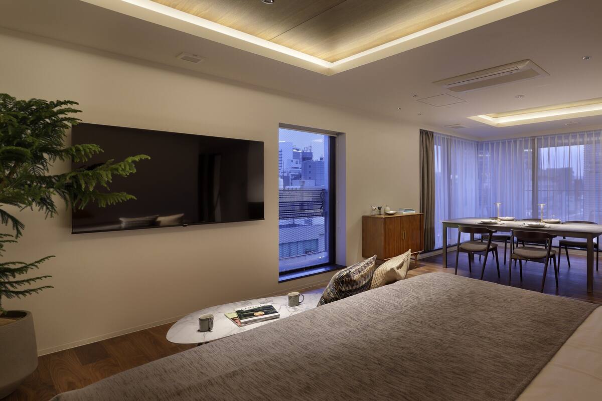 A modern hotel room with a large bed, a dining area, a TV mounted on the wall, and a view of the city through the window.