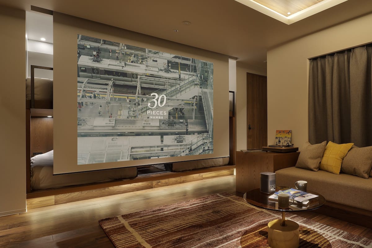 A modern living room with a large screen displaying a cityscape, a sofa with cushions, a coffee table with a mug, and a wooden floor.