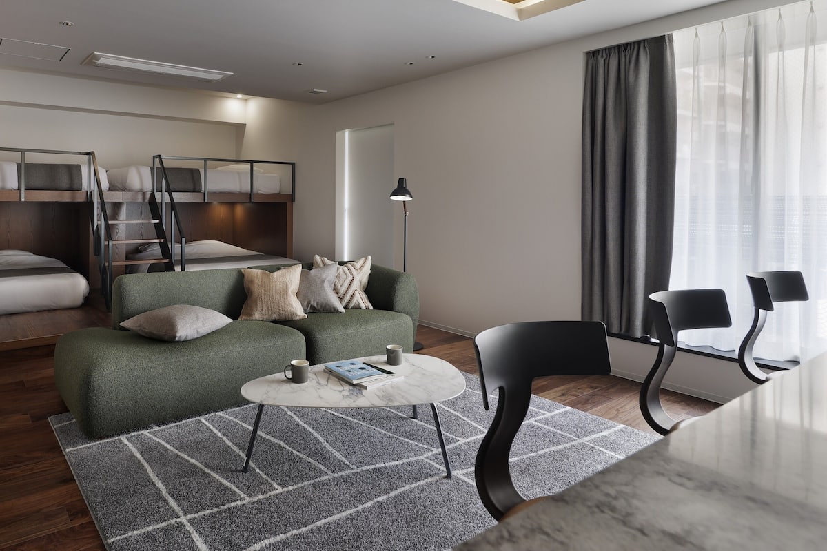 A modern hotel room with a bunk bed, a green sofa, a small coffee table, and a desk with two chairs.