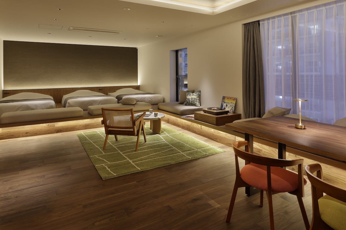 A modern hotel room with a large bed, a dining area, a seating area, and a large window with curtains.
