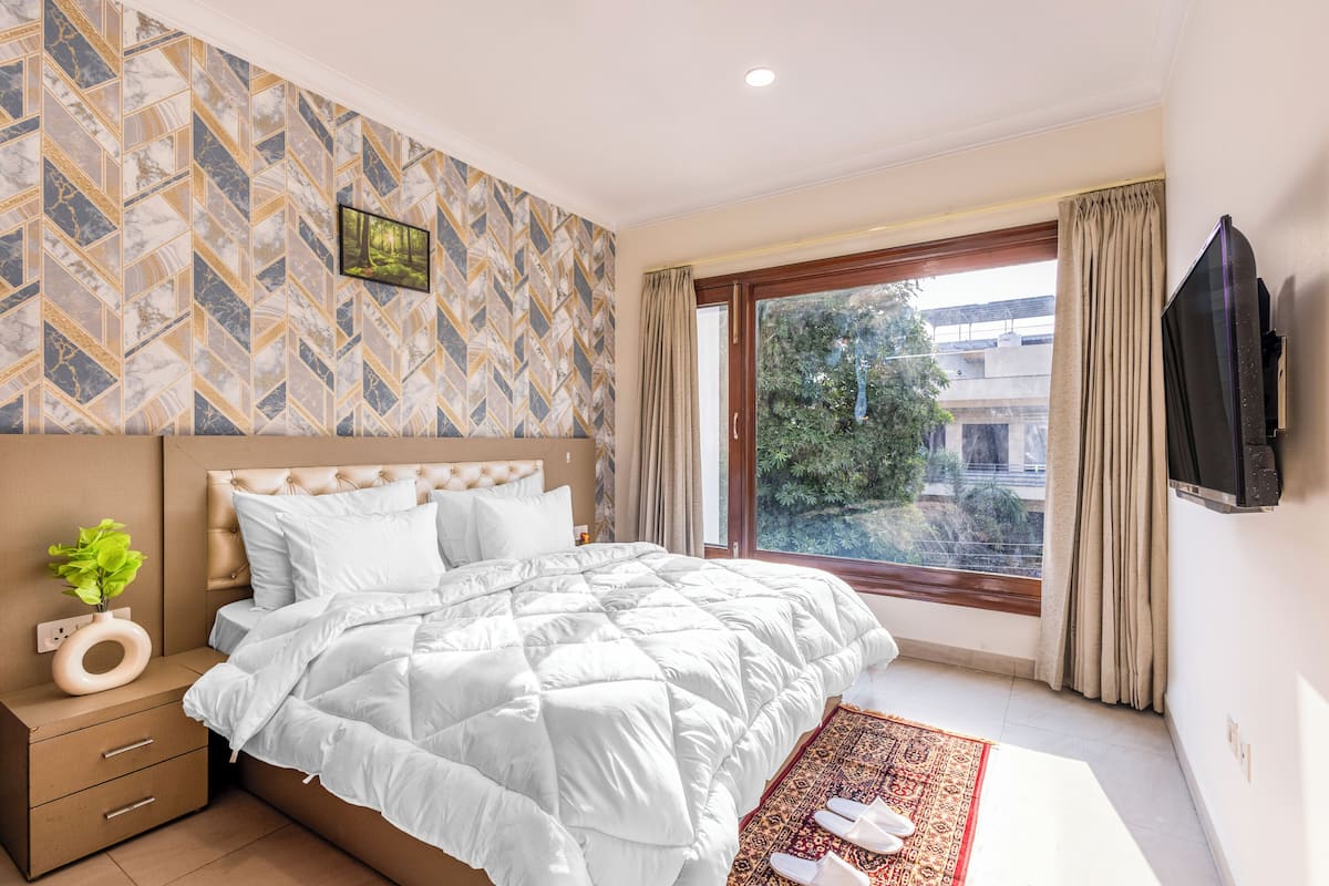 Een slaapkamer met een groot bed, een aan de muur bevestigde televisie, een raam met gordijnen en een kamer met een patroon op de muur.