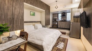 Ein modernes Hotelzimmer mit einem großen Bett, einem Esstisch mit Gläsern, einer Pflanze und einem durch eine offene Tür einsehbaren Küchenbereich.