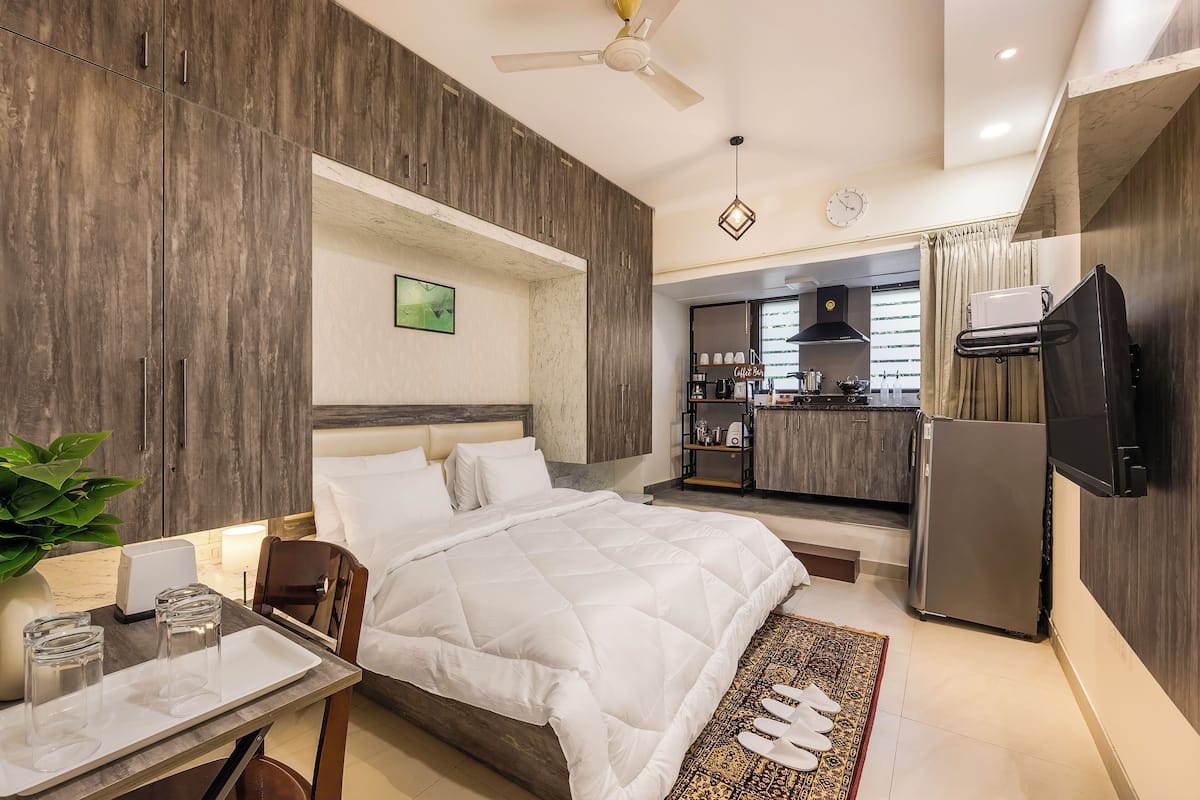 Een moderne hotelkamer met een groot bed, een eettafel met glazen, een plant en een keukenruimte zichtbaar door een open deur.