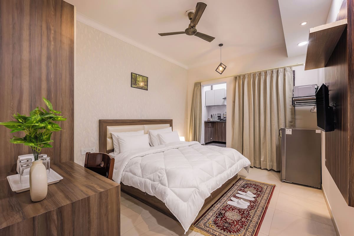 Een hotelkamer met een bed, een bureau, een plant, een plafondventilator en uitzicht op de keuken.