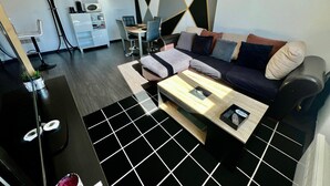 A modern living room with a sectional sofa, a coffee table, and a TV stand. There is a dining area with a table and chairs in the background. - One-Bedroom Apartment – Saint-Éloi, Poitiers (Poitiers)