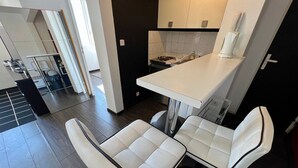 A modern kitchen with a white countertop, two white cushioned chairs, and a dark wooden floor. - One-Bedroom Apartment – Saint-Éloi, Poitiers (Poitiers)